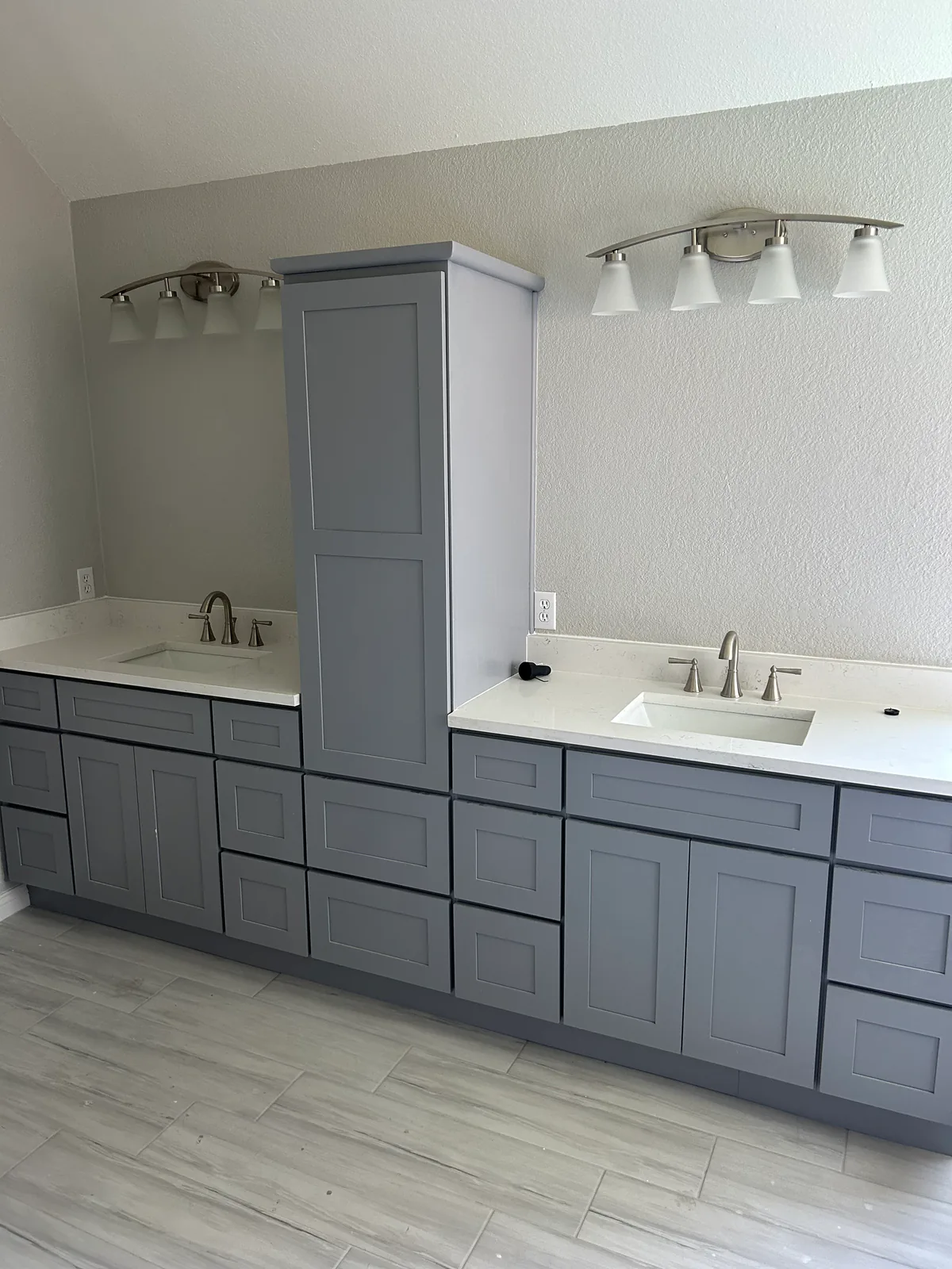 A finished bathroom remodel by AA Flooring & Design — grey shaker double vanity with white quartz countertops, undermount sinks and wood-look plank tile flooring